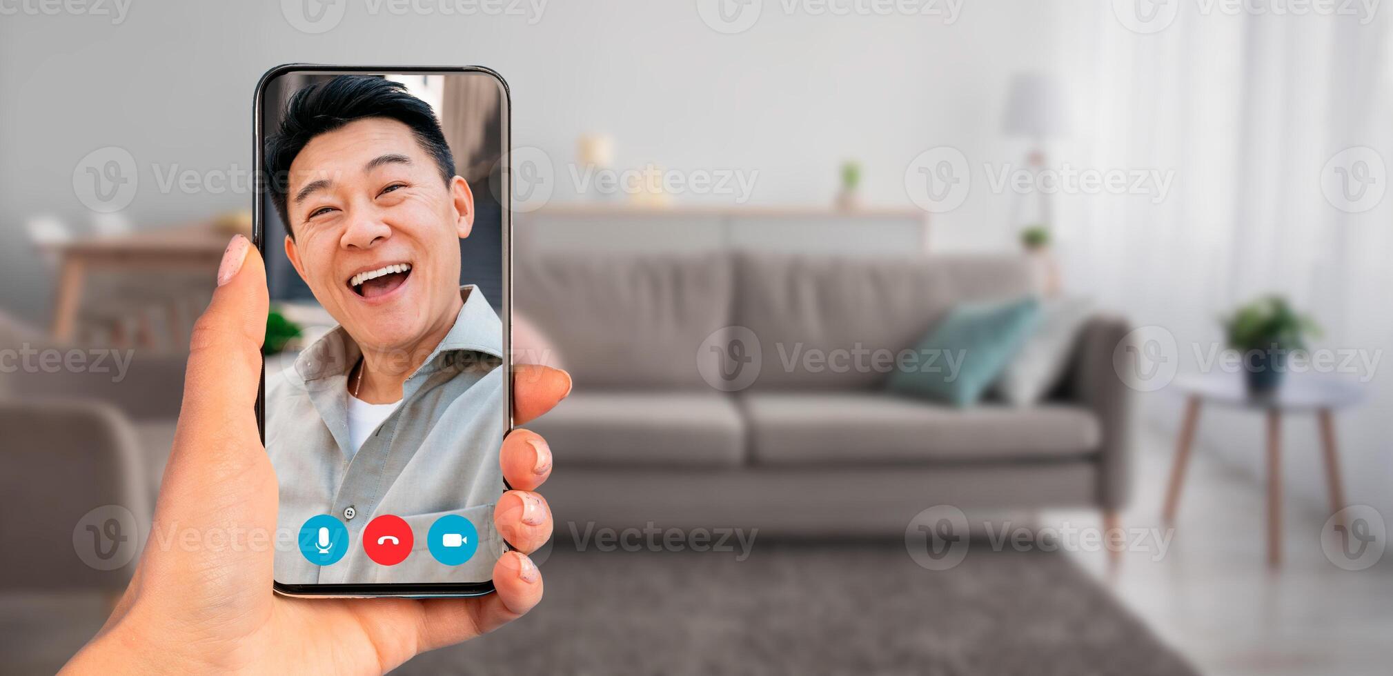 A person holding smartphone during a call with Asian man. The setting is a modern living room featuring comfortable furniture and soft lighting, perfect for virtual communication. photo