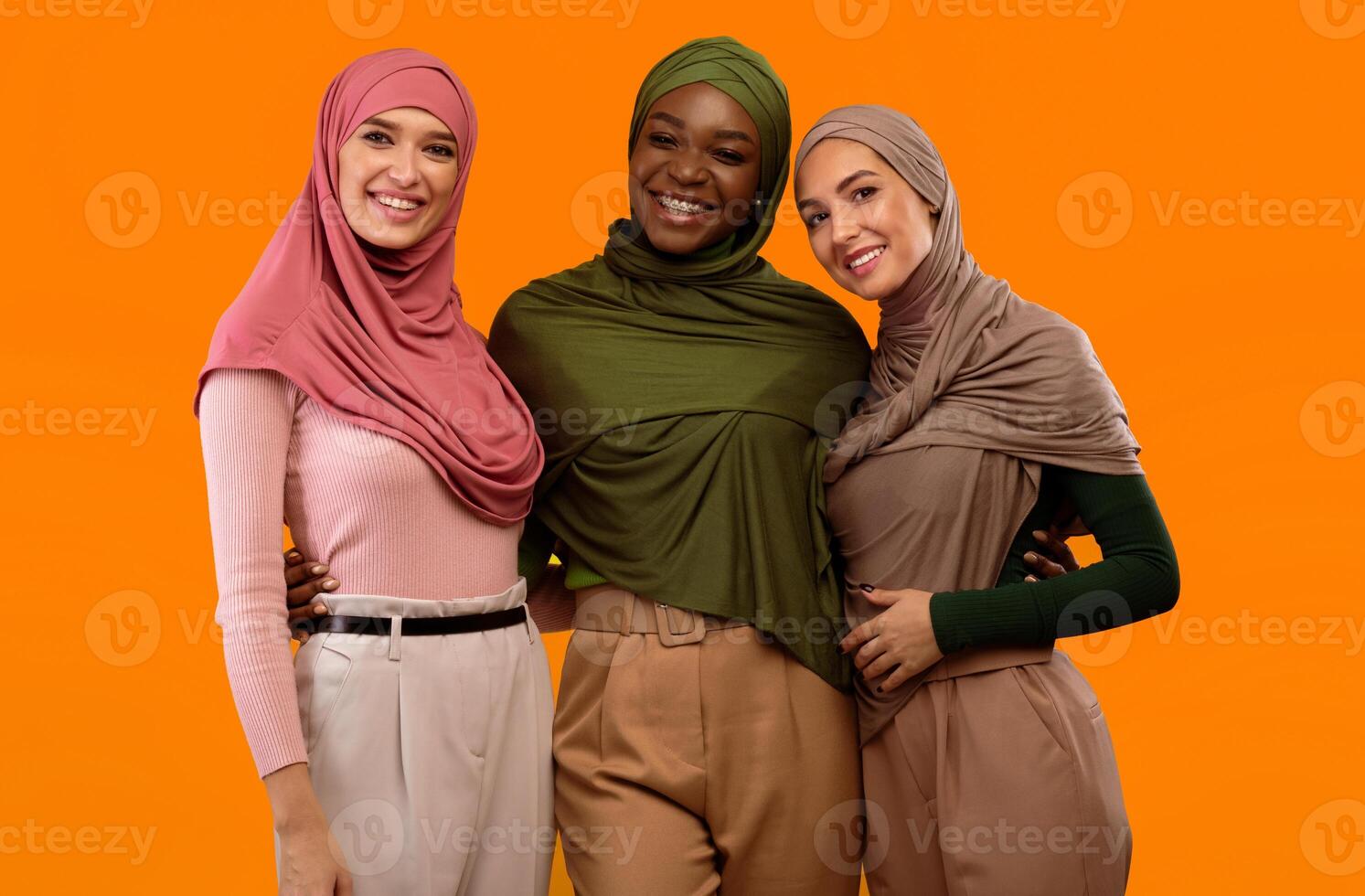Three Muslim Ladies In Hijab Headscarf Posing Standing Over Yellow Studio Background, Smiling To Camera. Cheerful Modern Islamic Ladies. Arabic Fashion And Female Frienship Concept photo
