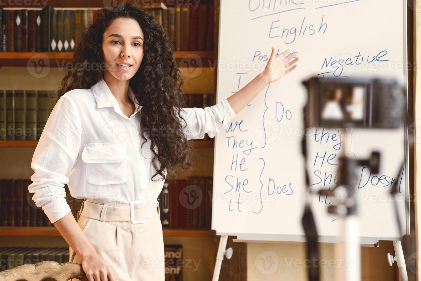 Online Lesson. Young female English language professor standing at whiteboard and teaching grammar rules, giving remote lecture. Woman recording blog for her internet channel at camera photo