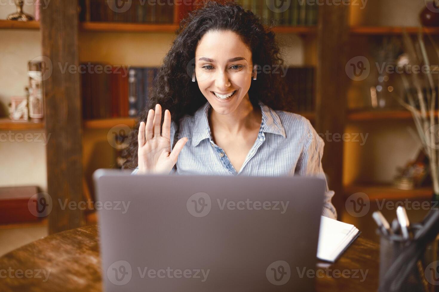 Online Call. Portrait of smiling woman sitting at desk, making virtual conference on laptop, waving with hand to webcam. Communication Concept. Woman chatting using computer, wearing earbuds photo
