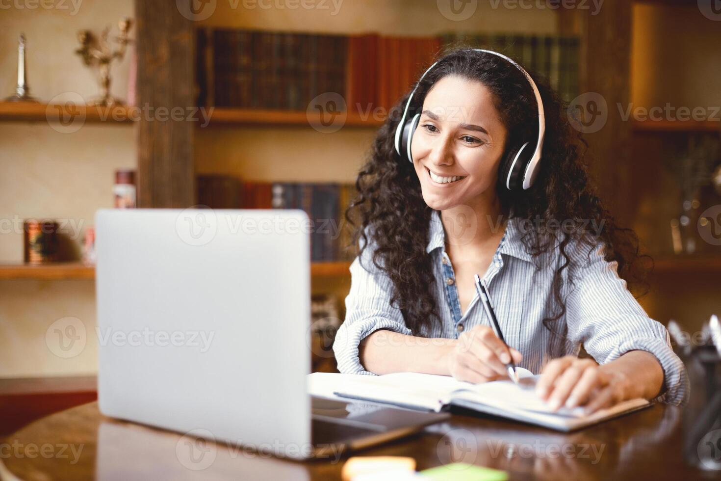 Home Office. Portrait of smiling young lady in wireless headphones sitting at desk, writing in notebook and using computer. Woman watching online tutorial, webinar or seminar, studying at home photo