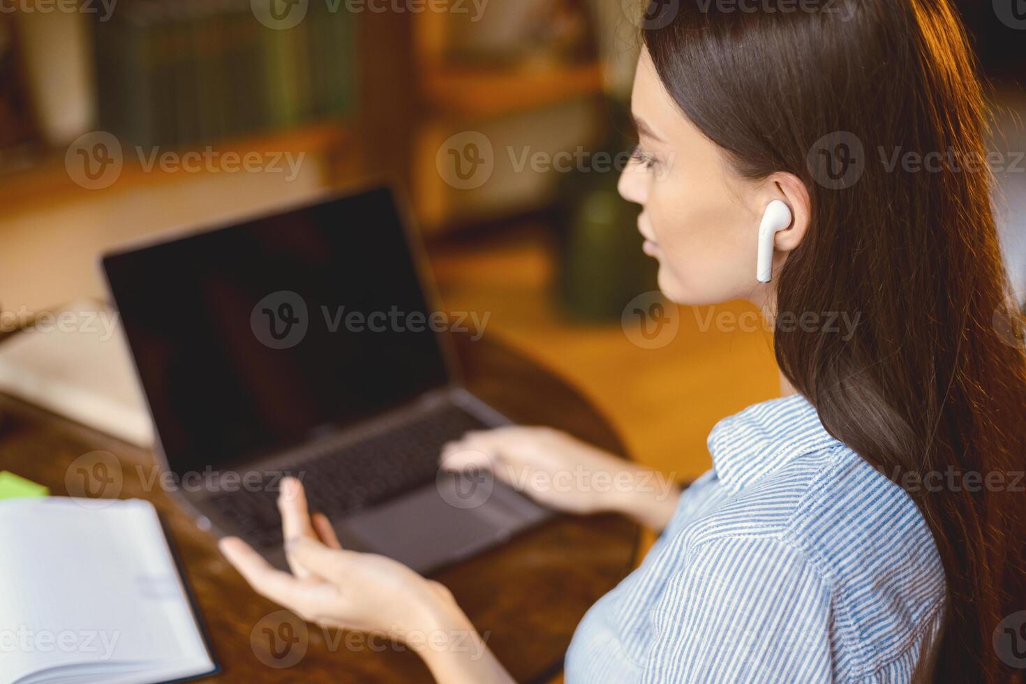 Virtual Meeting Concept. Back over the shoulder view of young woman in wireless earphones sitting at desk, using laptop with black screen for online videocall conference, selective focus photo