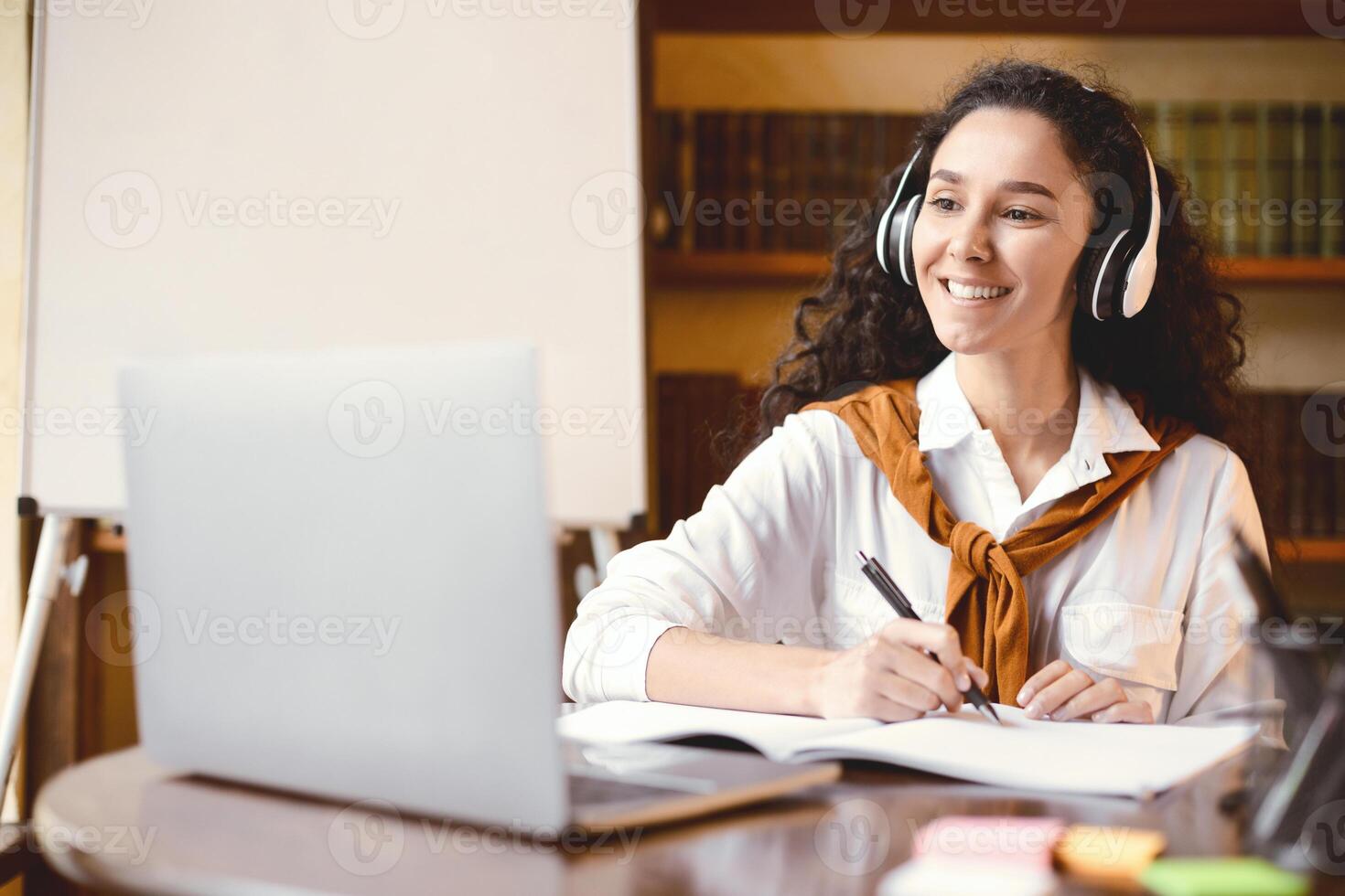 E-learning Concept. Positive cheerful young curly woman watching virtual lecture, looking at laptop, writing with pen and taking notes in her notebook, sitting at table. Education during lockdown photo