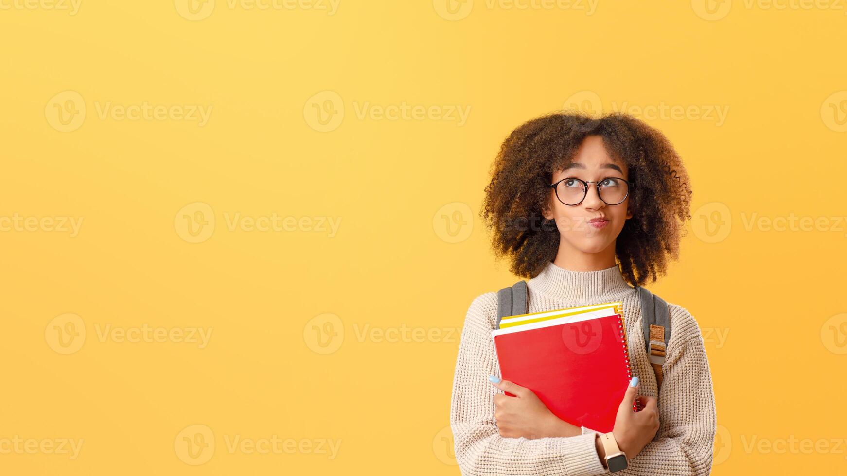 Think and make decisions. Pensive african american lady in glasses with smart watch and backpack holding notebooks and looks up at empty space, isolated on yellow background, studio shot, cut out photo