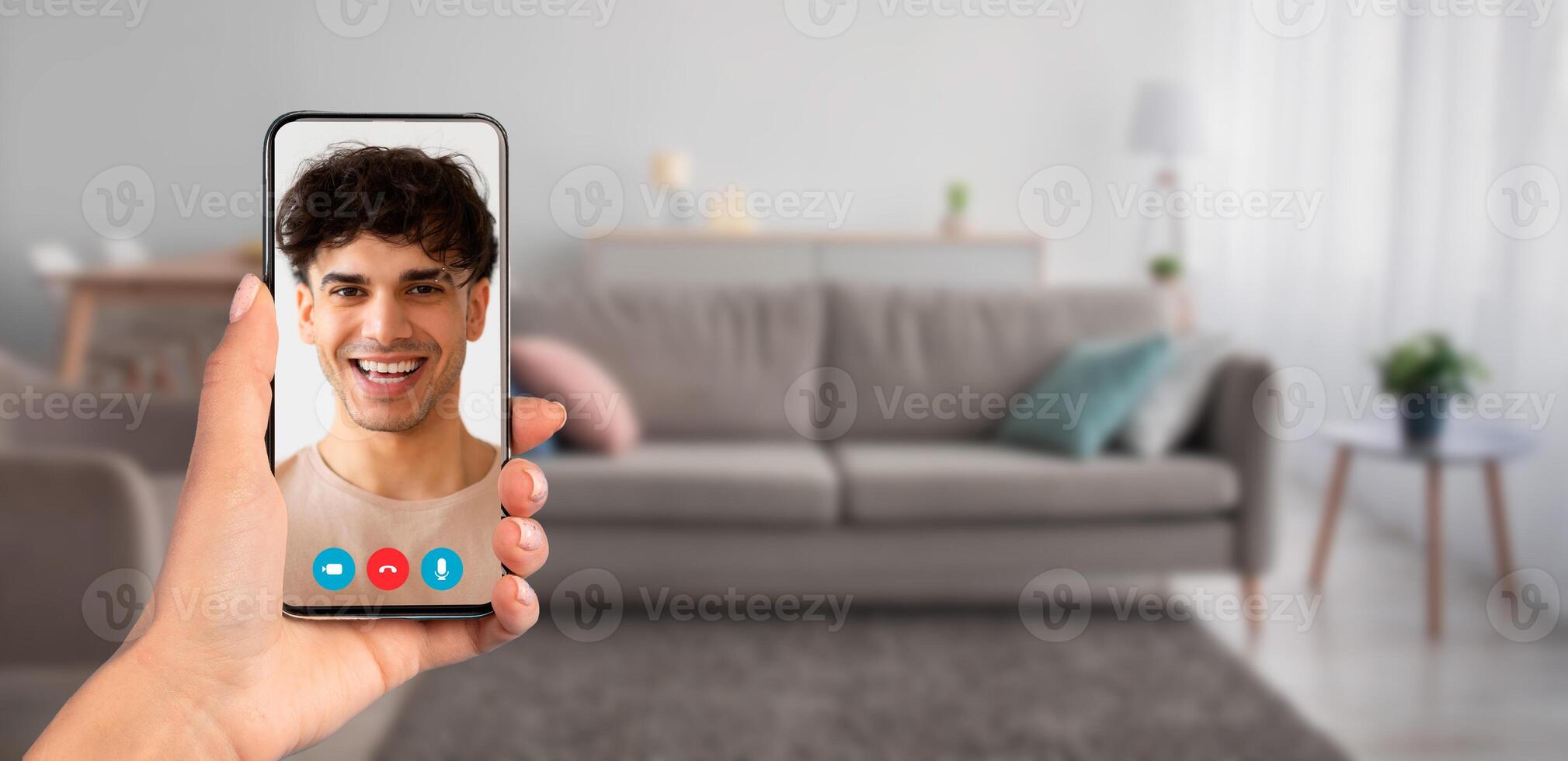 A person holding a smartphone during a call at home. The cozy living room features a sofa and soft lighting, creating a comfortable atmosphere for virtual interaction. photo