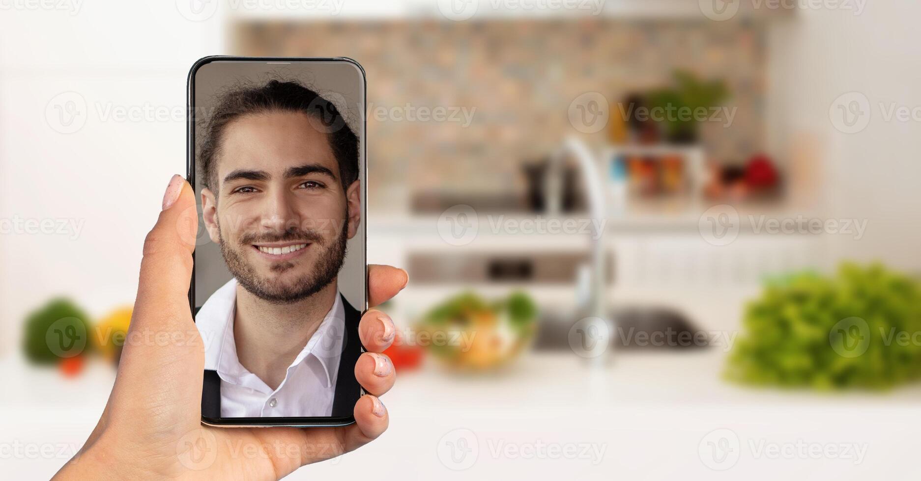 A person holds a smartphone displaying a smiling man during a call. The background features a modern kitchen with fresh vegetables and a bright atmosphere, indicating a casual home setting. photo