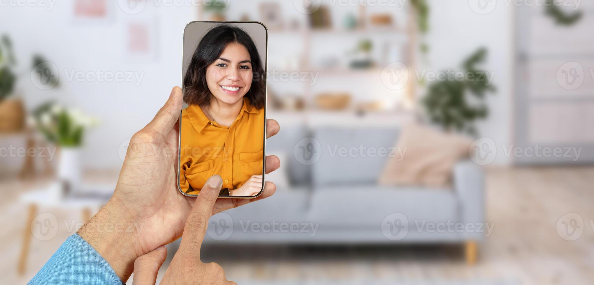 A person engages in an online call using a smartphone, smiling at the screen. The cozy home environment features a sofa and plants, creating a friendly atmosphere for communication. photo