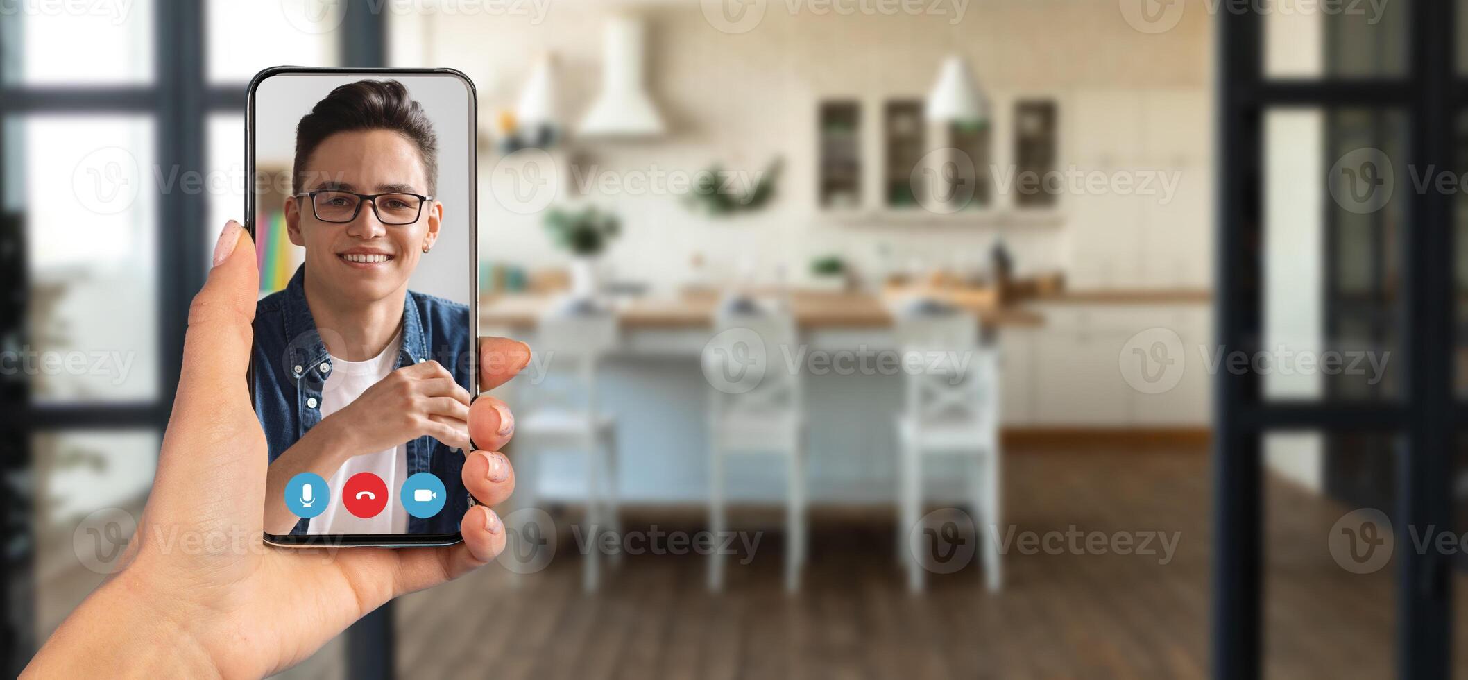 A person is engaged in an online call using a smartphone in a stylish home kitchen. The setting features a bright, open space with modern furniture and decor, creating a warm ambiance. photo