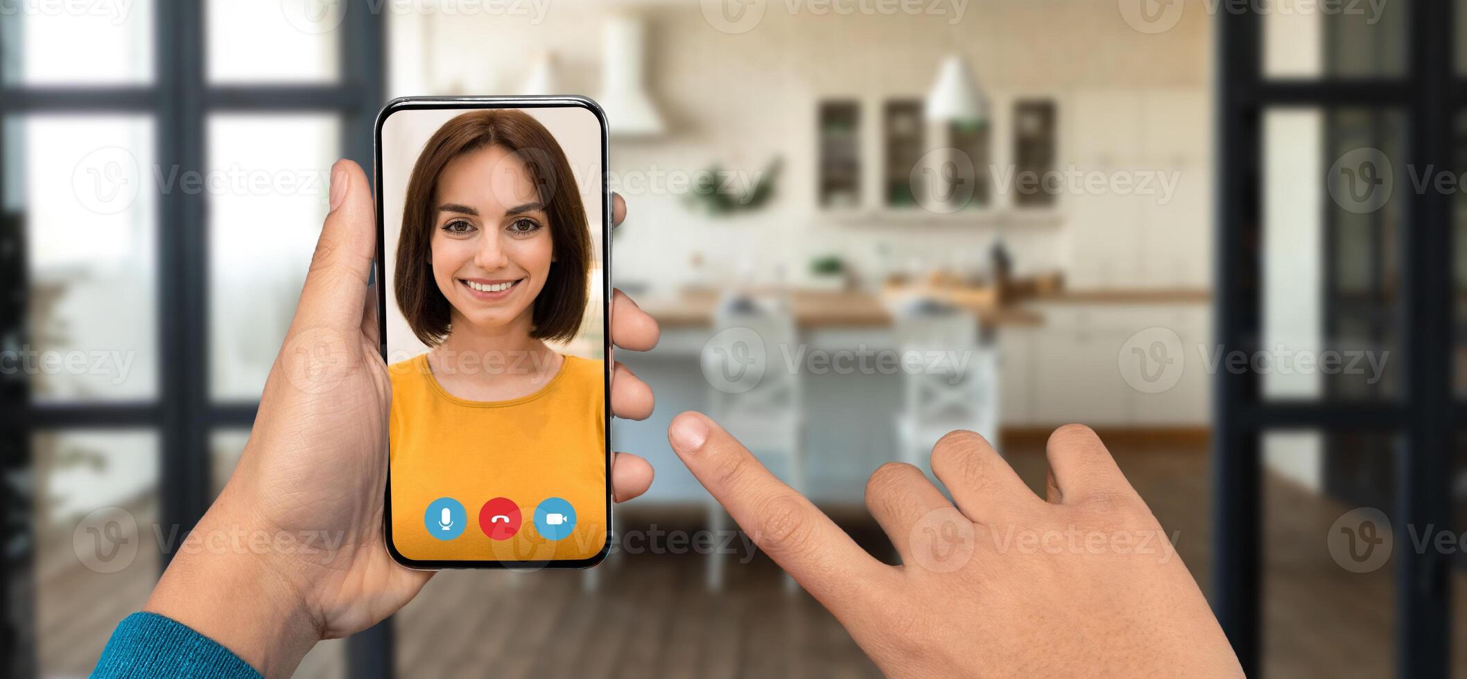 A person is holding a smartphone to engage in a lively call while in their cozy home. The background features a well-lit kitchen setting, enhancing the atmosphere of connection. photo