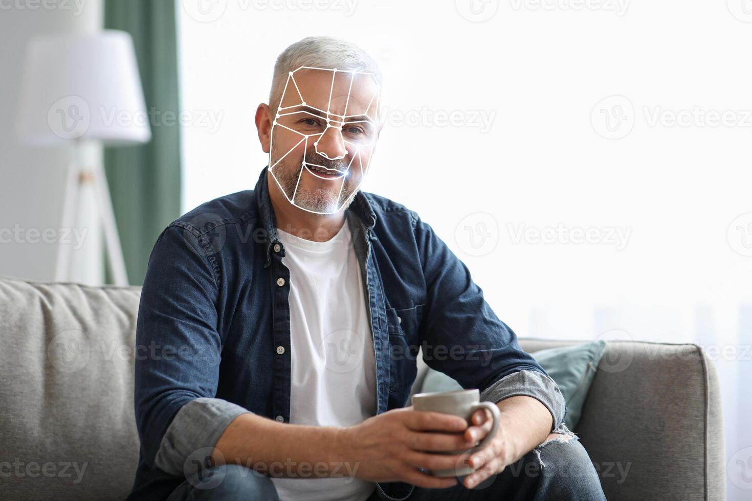 A man sits comfortably on a couch holding a mug, engaged in facial recognition technology. The bright, serene room enhances the sense of modern living and adaptation to new technology. photo