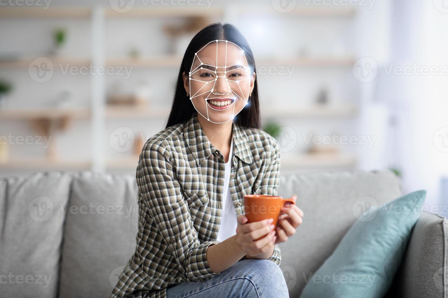 Asian woman sits comfortably on a sofa, holding a cup while engaged in facial recognition technology. This scene highlights the intersection of modern technology and personal identification at home. photo