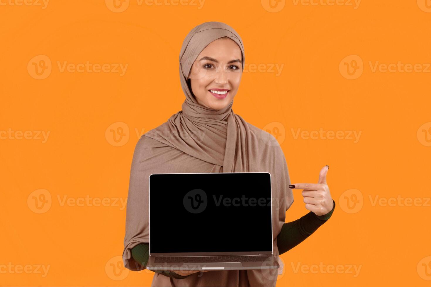 Great Website. Muslim Lady Pointing At Laptop Computer Empty Screen Recommending Internet Service Or Blog Standing Posing On Yellow Studio Background. Mockup photo