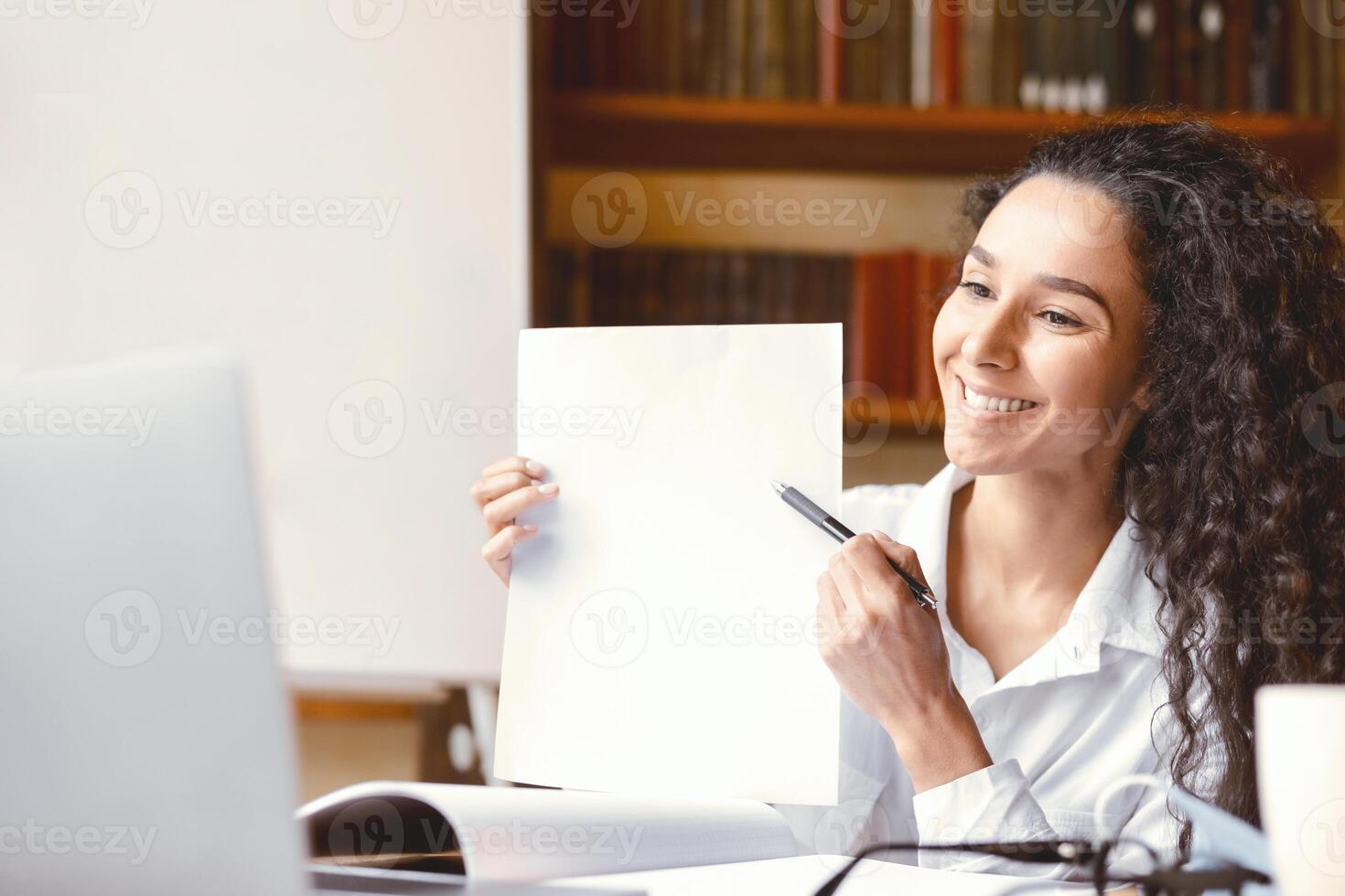 Smiling female manager having videocall on laptop, giving virtual presentation or online lesson to students. Woman showing document contract to webcam, pointing to blank empty sheet of paper photo