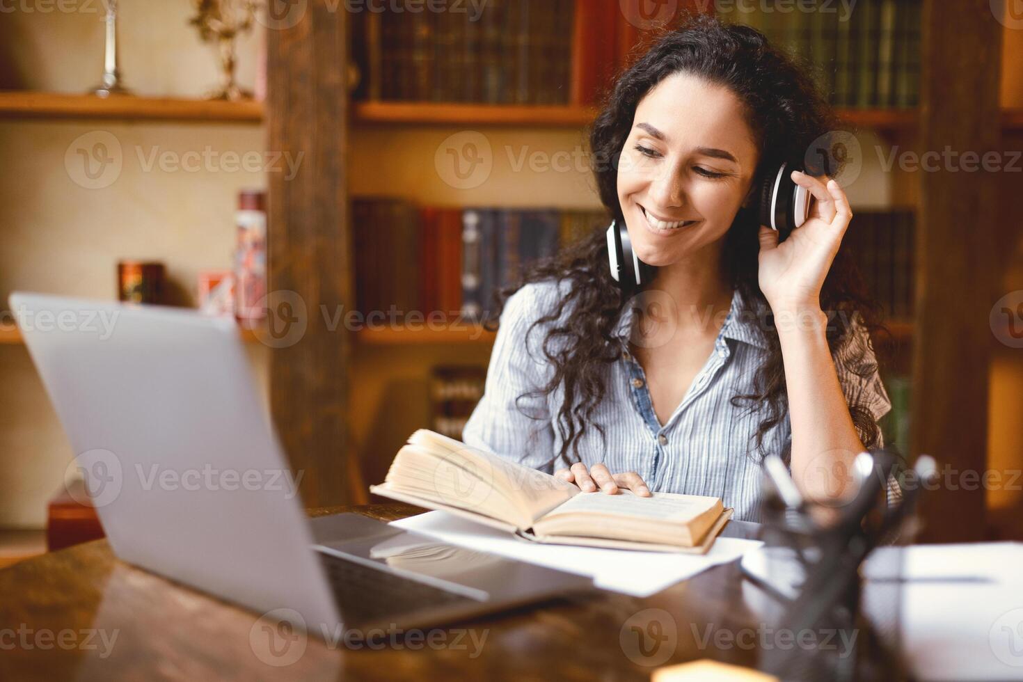 Portrait of smiling lady touching wireless headsetphones, sitting at table, using computer and reading book. Happy female watching online tutorial, lecture or class, studying online at home. Elearning photo