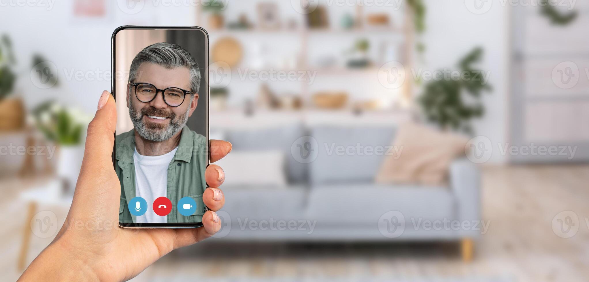 A person holds a smartphone displaying a smiling man on a call. The background reveals a cozy living room environment with a sofa and decor, indicating a relaxed setting for communication. photo