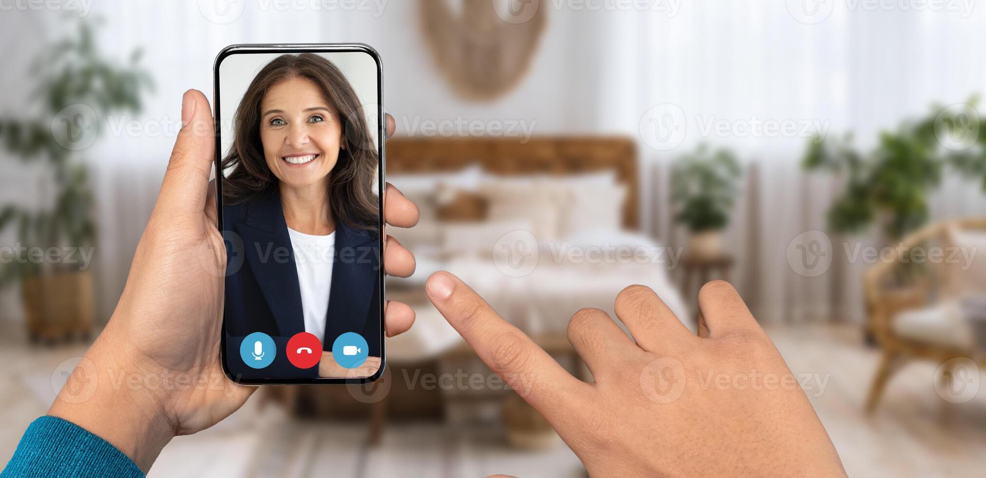 A person is holding a smartphone displaying a smiling face during an online conversation at home. The cozy room features a bed and potted plants, emphasizing a relaxed atmosphere. photo