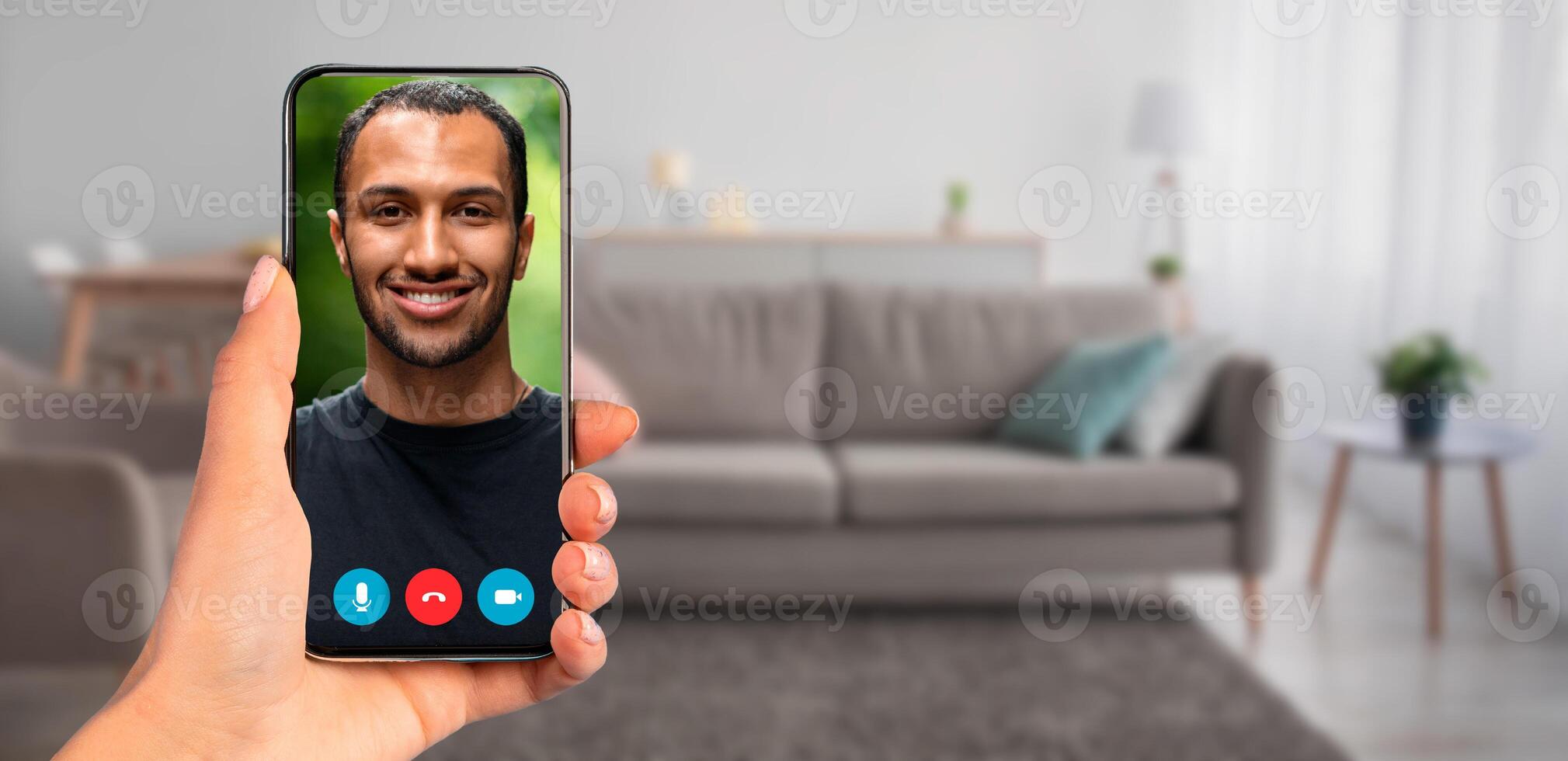 A person holding a smartphone that displays a call with handsome guy. The background features a cozy living room with a couch and natural light, creating a relaxed atmosphere for communication. photo