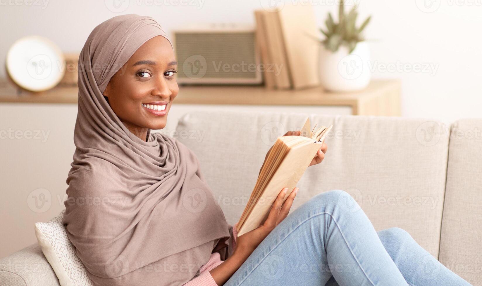 Happy black lady in traditional hijab sitting on comfy sofa with book at home, copy space. Beautiful Muslim lady in Arabic headscarf relaxing on couch, reading interesting story, indoors photo