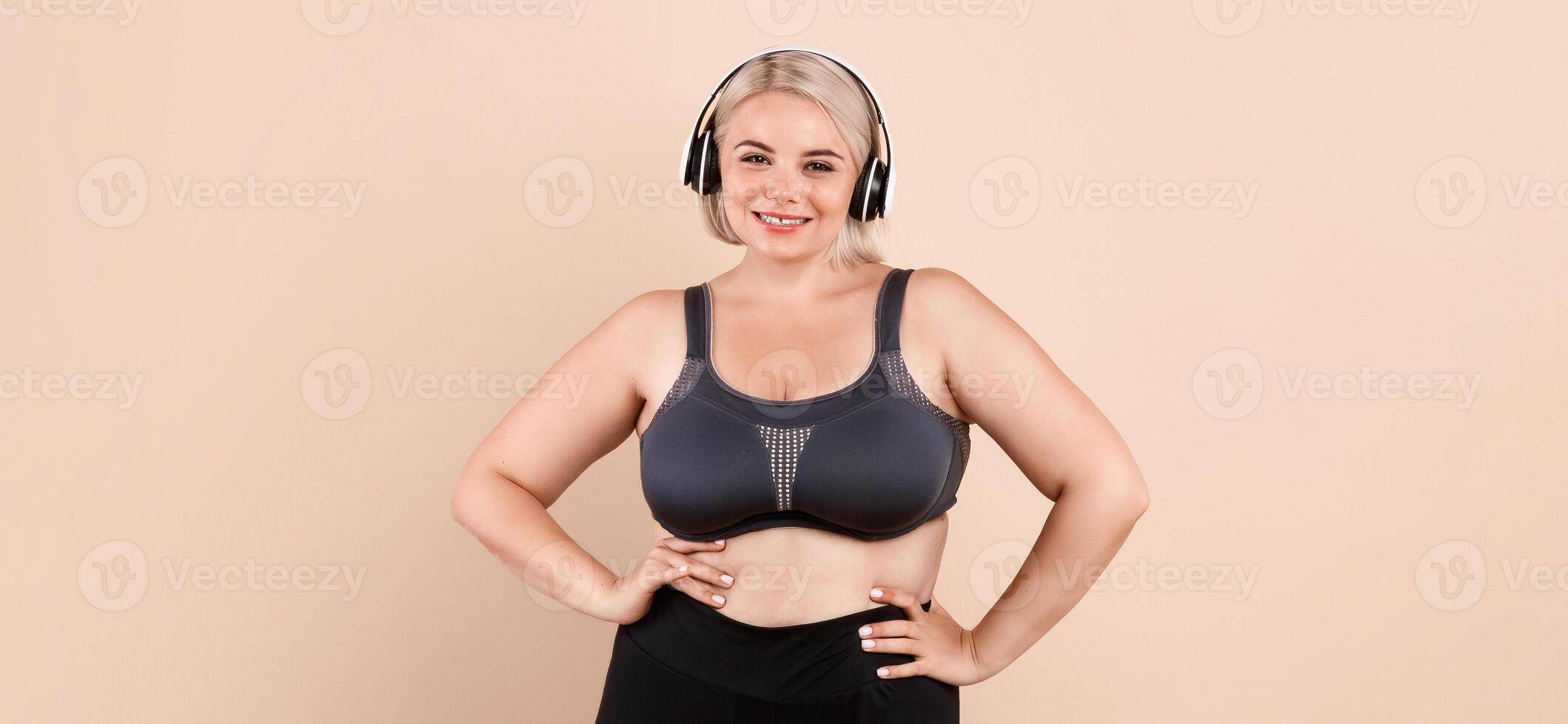 Weightloss Concept. Happy Plus Size Woman In Sporswear Posing Over Beige Studio Background, Curvy Young Lady Wearing Wireless Headphones Ready For Training, Smiling At Camera, Copy Space photo