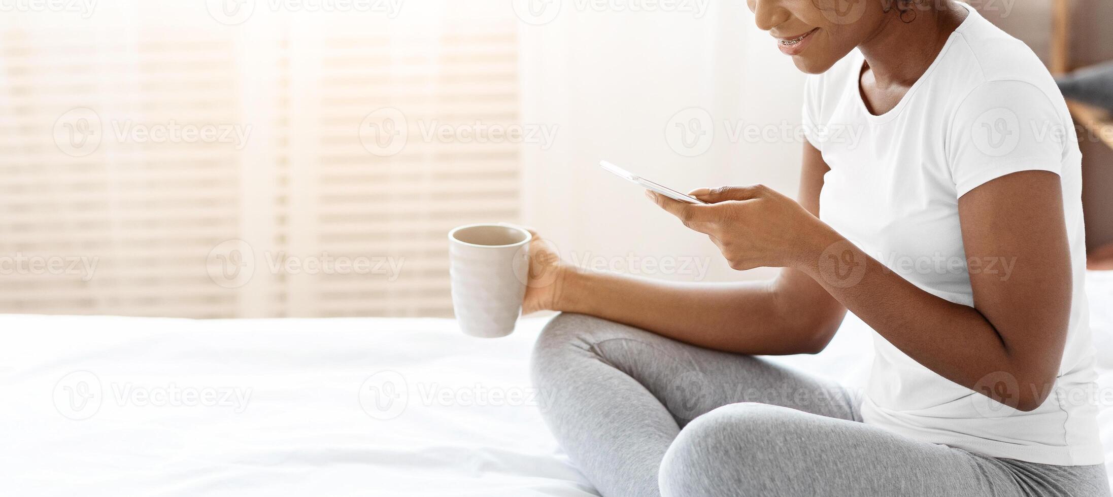 Smiling african american young lady in pajamas drinking morning tea or coffee, using mobile phone, checking social media or reading messages, sitting on bed at home, side view, copy space photo