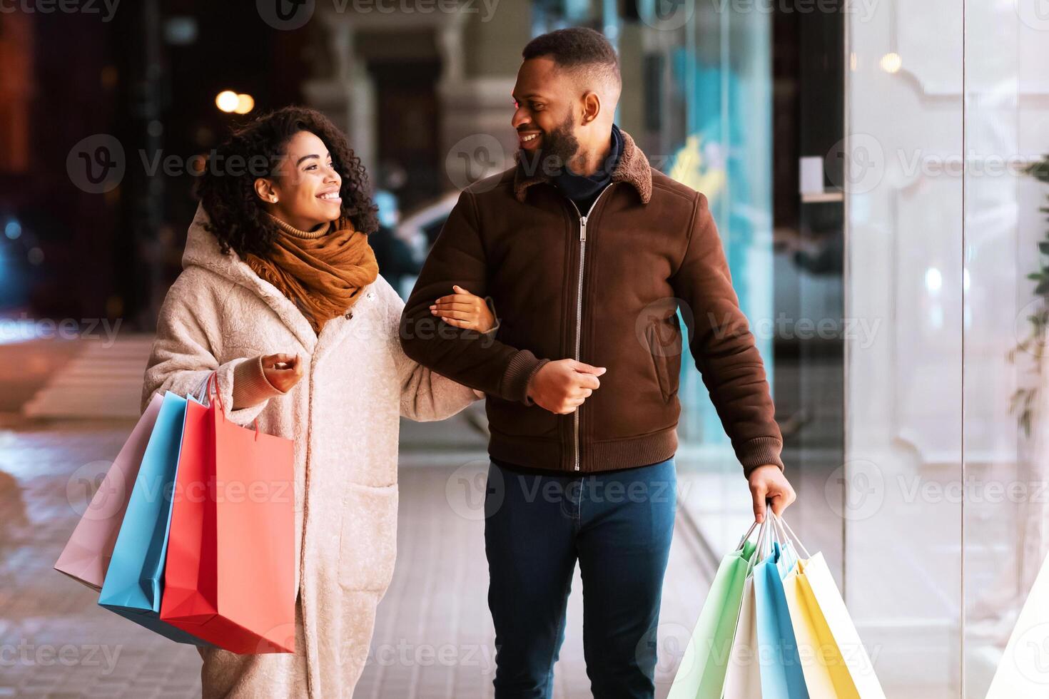 Family Shopping Concept. Full length portrait of happy black african american couple walking down the street in the evening, holding shopper bags, looking at each other and smiling. Consumerism photo