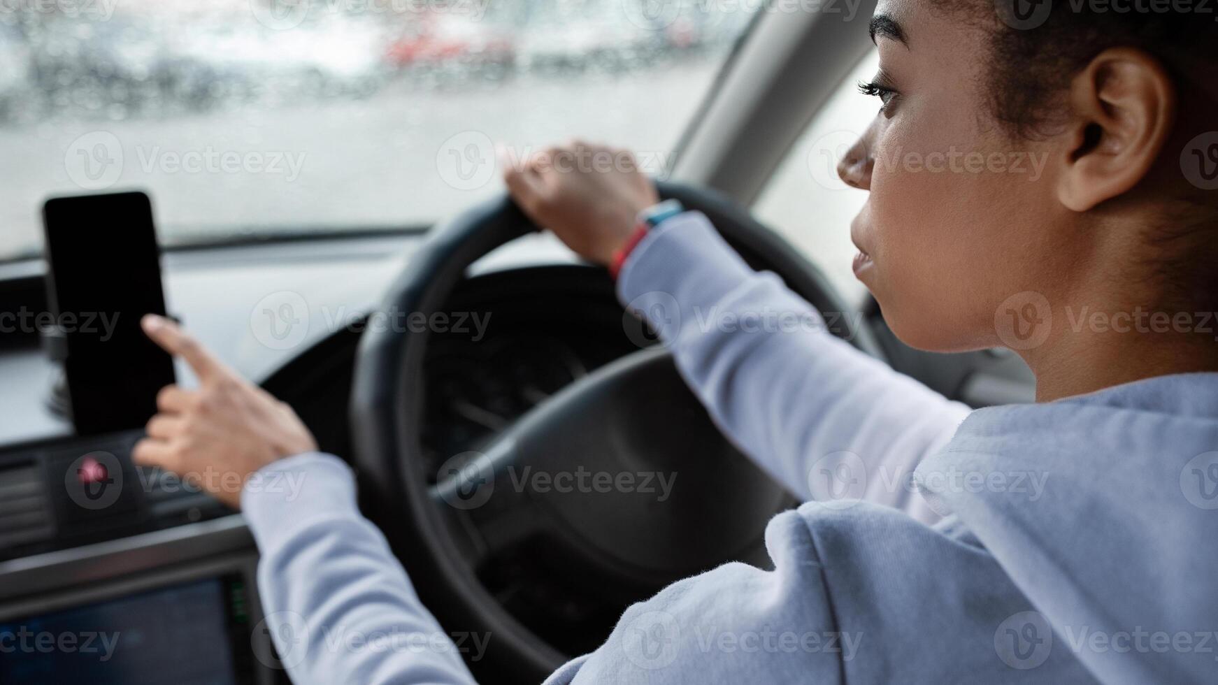 Make route without traffic jams on way to work. Serious young african american woman drives car and use smartphone with empty screen and online map in rainy weather, cropped, side view, copy space photo