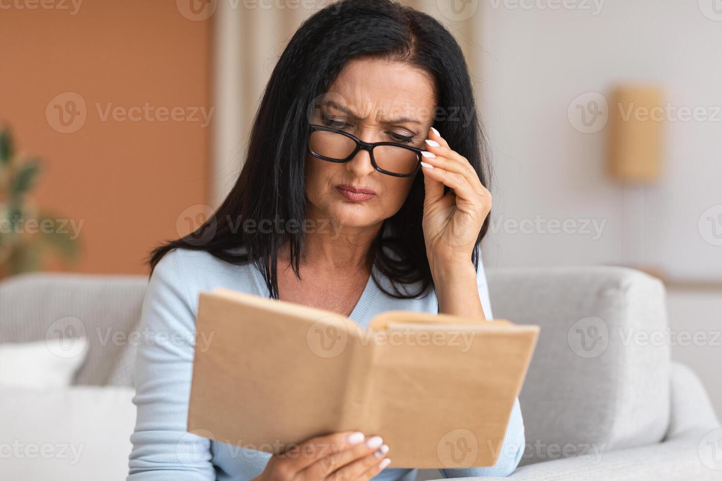 Portrait of mature woman touching glasses trying to read book, having difficulties seeing text because of vision problems. Negative emotion facial expression feelings reaction, health issues photo