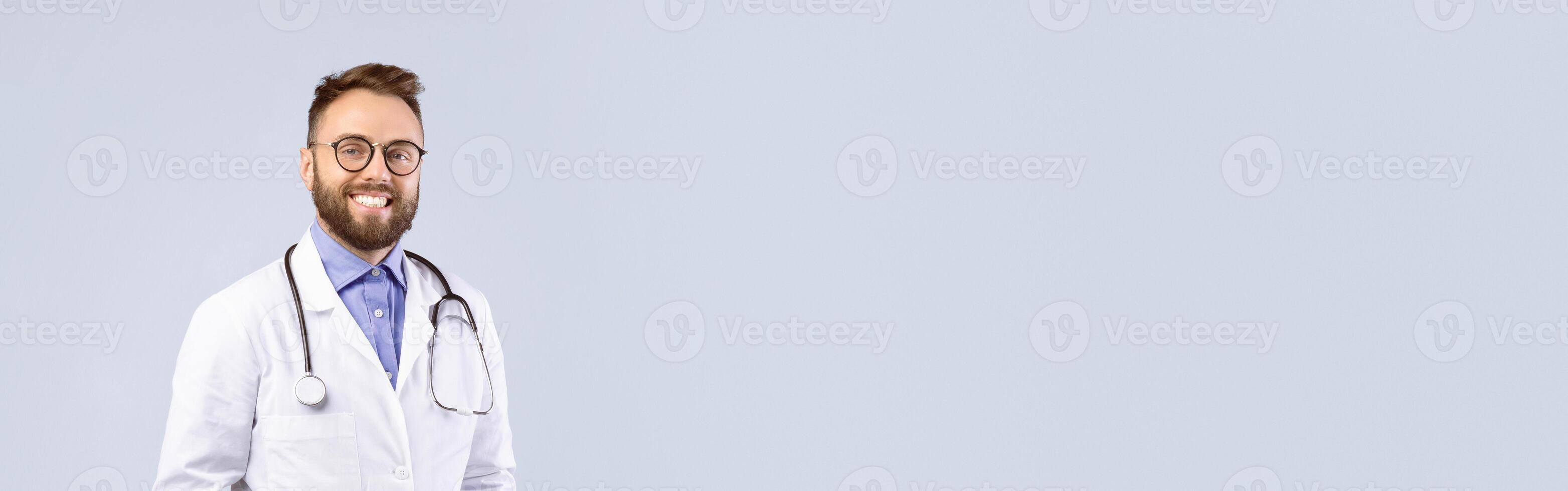 Confident Caucasian male doctor in white uniform smiling and posing over grey studio background. Medical practitioner or healthcare professional with hands in pockets looking at camera photo