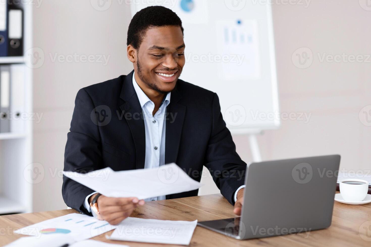 Happy black businessman in stylish suit holding documents and looking at laptop screen, checking reports in modern office, satisfied by results of his company, working on computer, copy space photo
