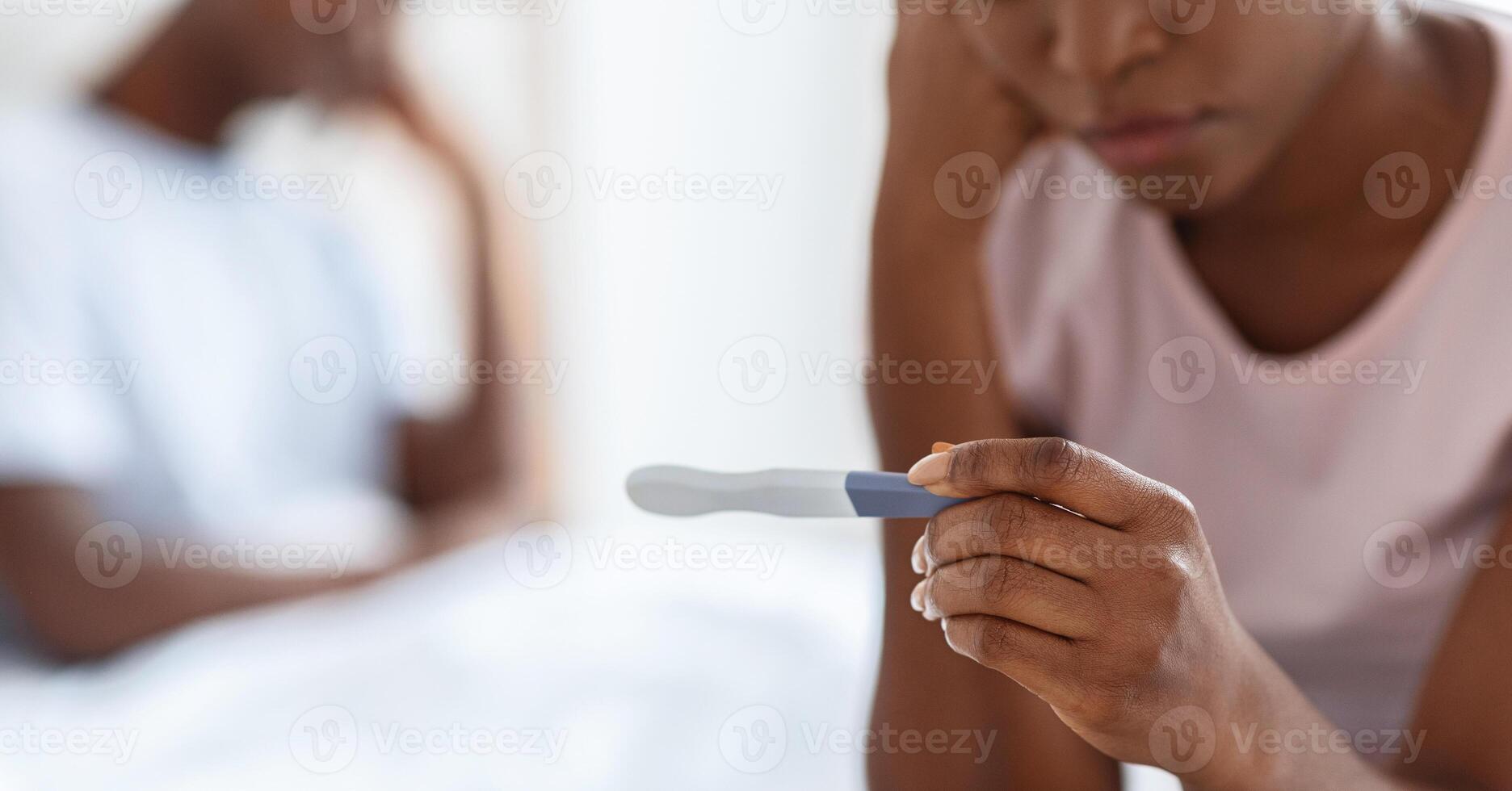 Unrecognizable black woman sitting on bed and holding positive pregnancy test, african american man in bed on background, unintended pregnancy concept, cropped, panorama. Selective focus on test photo
