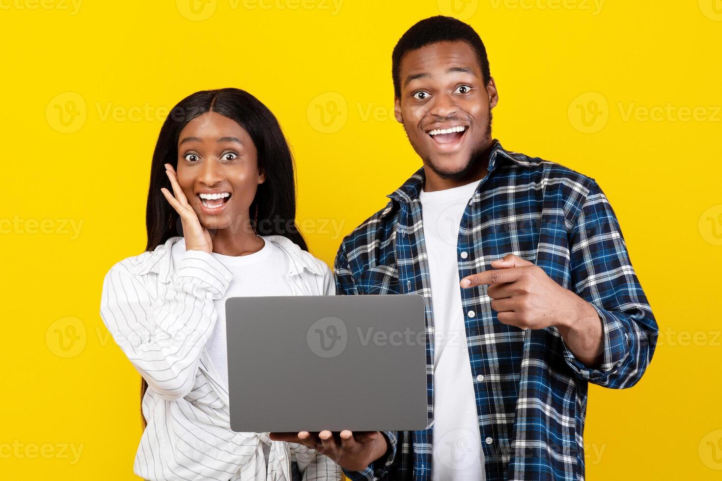 Amazing offer, good site and celebrating victory. Surprised cheerful millennial african american husband and wife hold laptop, man points finger at gadget, isolated on yellow background, studio shot photo
