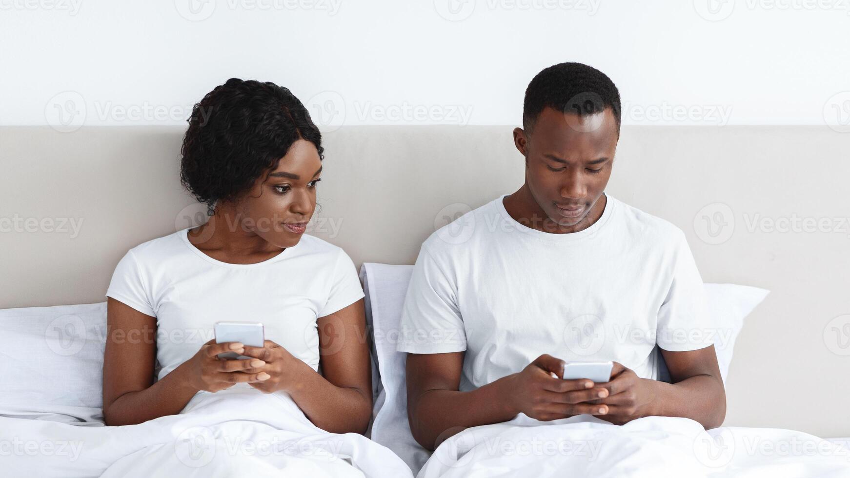Young black woman eavesdropping on her boyfriend using mobile phone, looking at his smartphone screen. African american couple sitting in bed and using mobile phones. Distrust in relationships photo