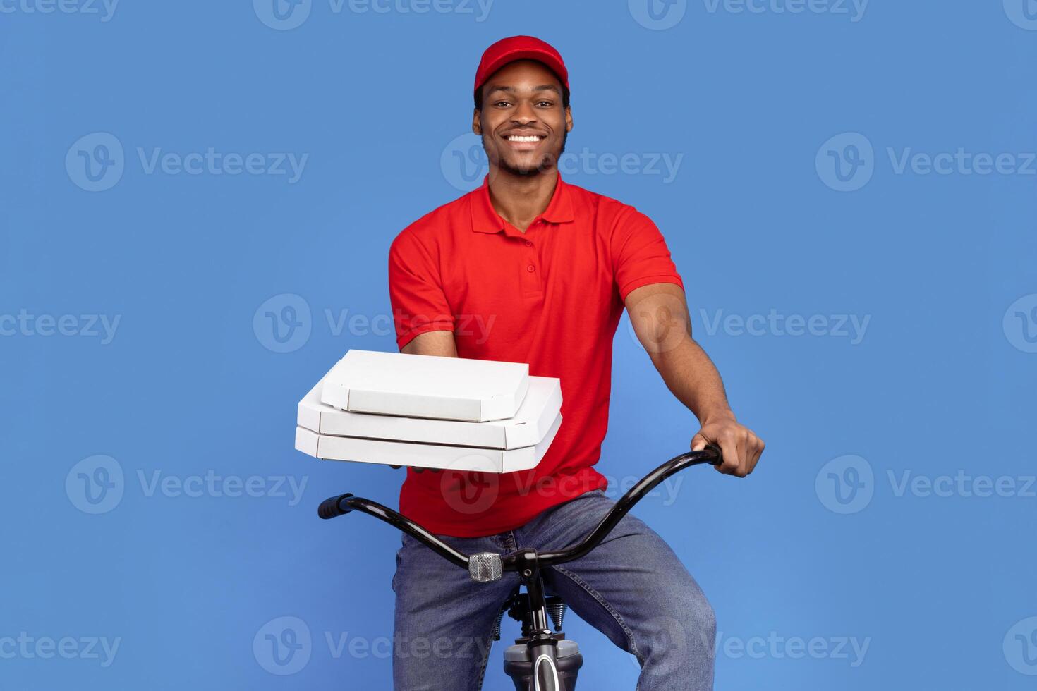 Best Fast Food Delivery. Portrait of smiling african courier wearing red cap and uniform holding pizza boxes from restaurant and riding bicycle, isolated on blue studio background photo