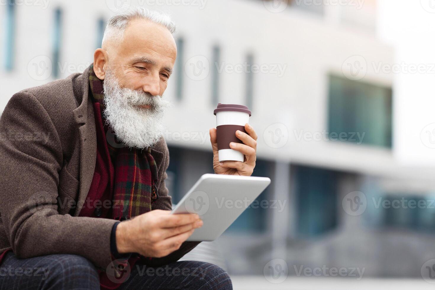 Grey-haired elderly man in warm outfit using digital tablet and drinking coffee to go while sitting on the street, enjoying his free time outdoors, reading book or checking social media, copy space photo