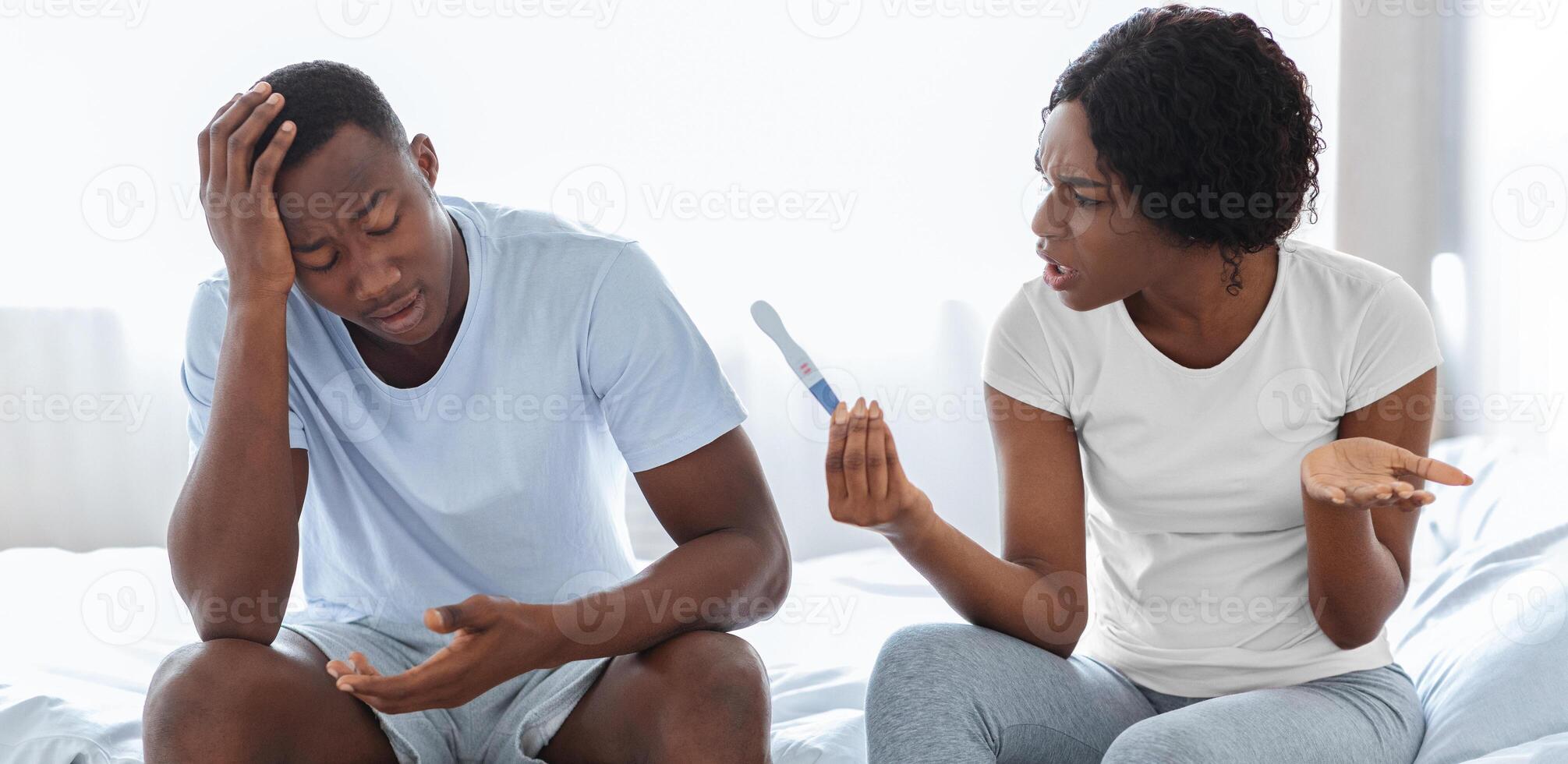 Unintended pregnancy and relationships. Angry black woman with pregnancy test yelling at her shocked confused boyfriend touching his head, african american couple sitting on bed at home photo