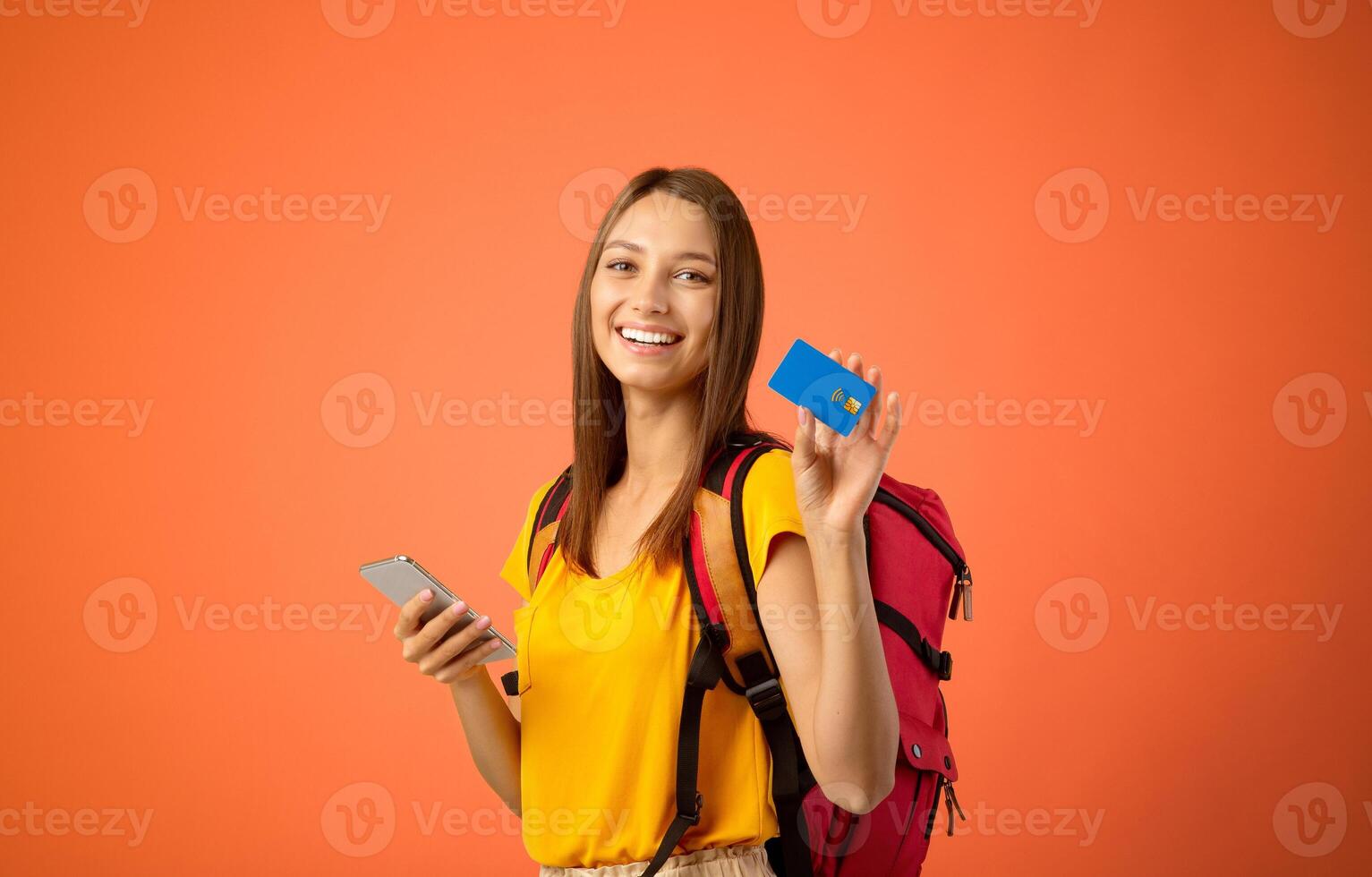 Happy woman backpacker with smartphone and credit card over yellow studio background, copy space. Cheerful young woman holding credit card and using mobile phone, shopping online while travelling photo