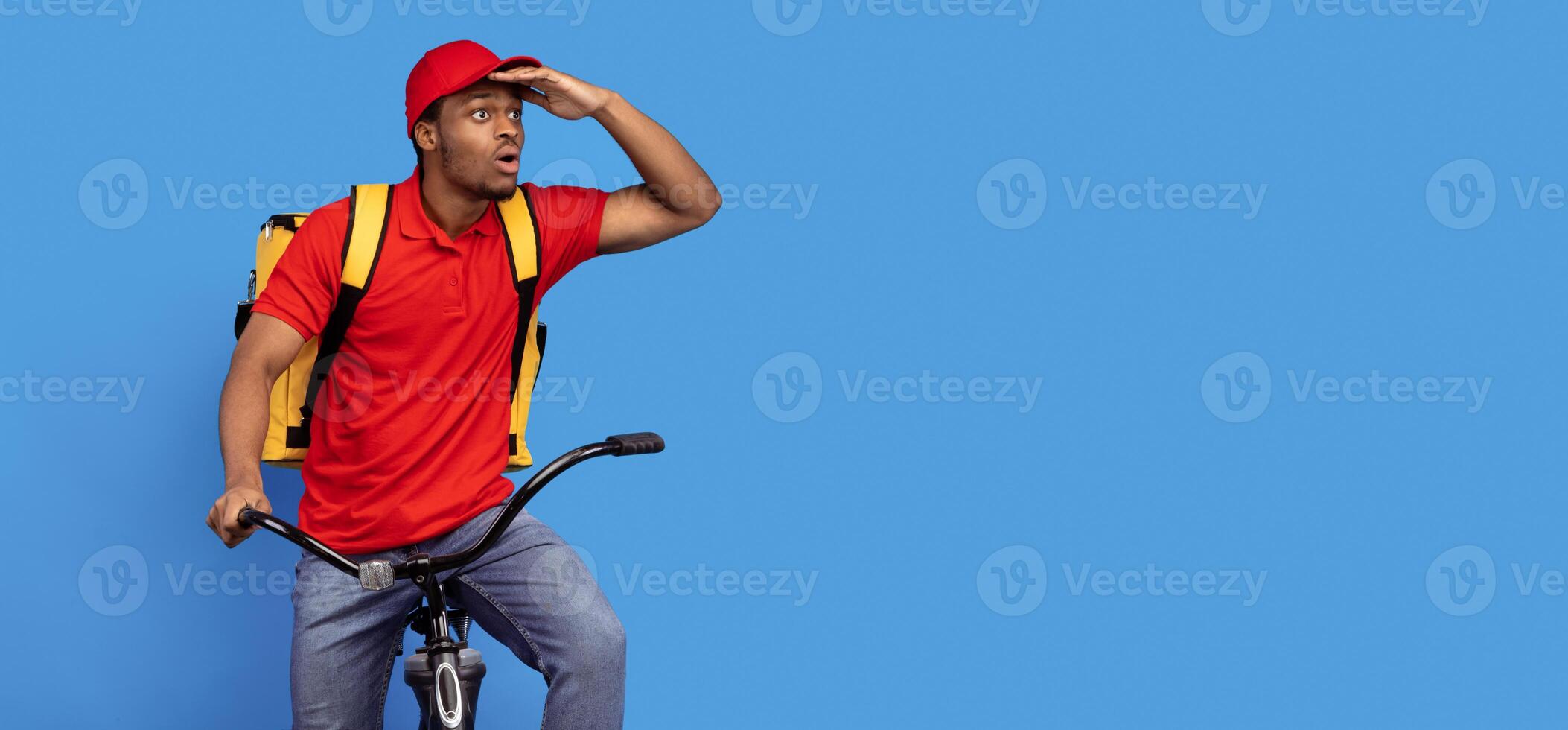 Fast Delivery Service. Emotional surprised black courier guy in red uniform riding bicycle, wearing thermo backpack bag and looking at copy space isolated on blue studio background, banner, panorama photo