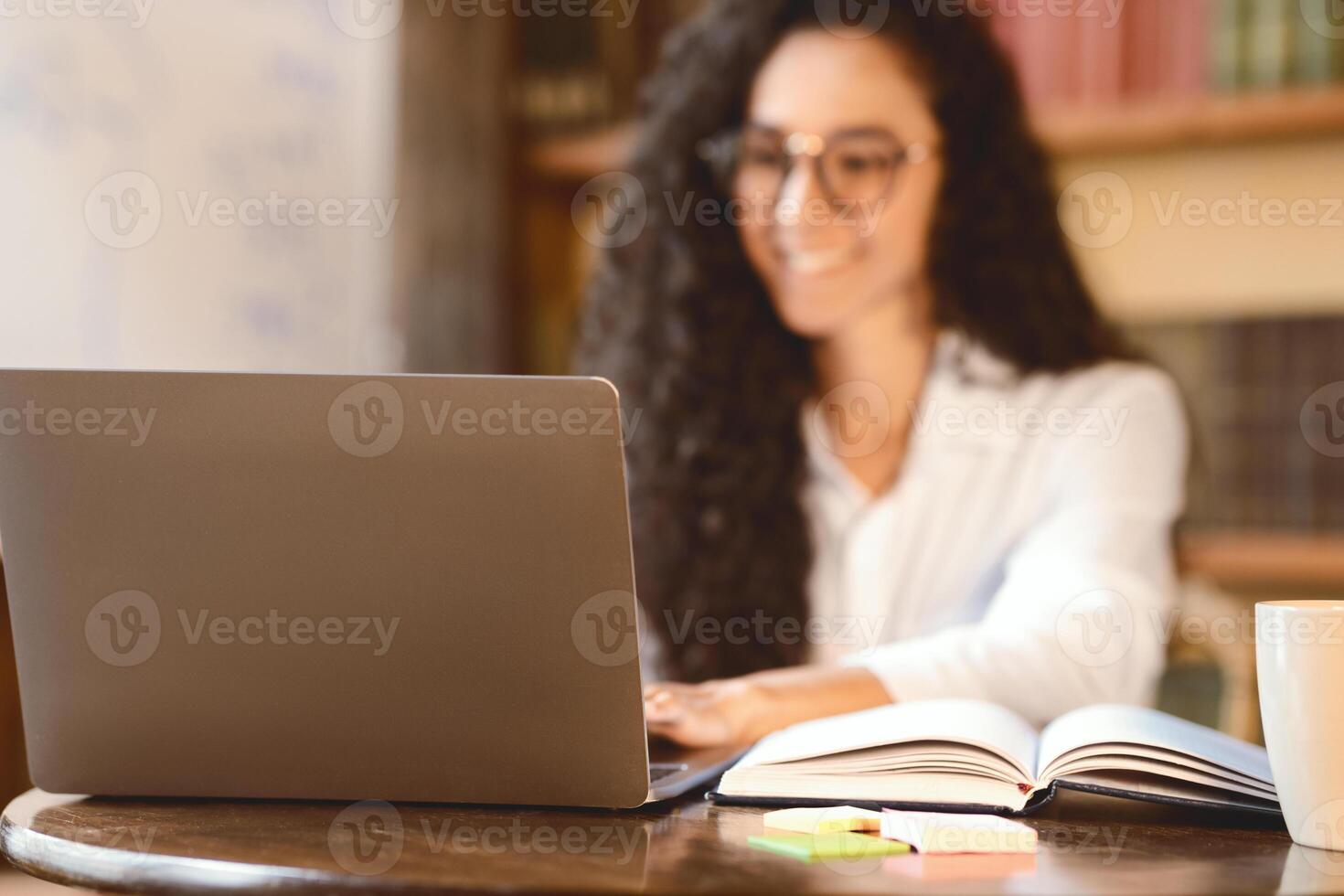 Workspace. Selective focus on notebook on the table, smiling woman in eyeglasses sitting at desk, using laptop in the blurred background. E-learning, remote work, blogger or journalist writing article photo