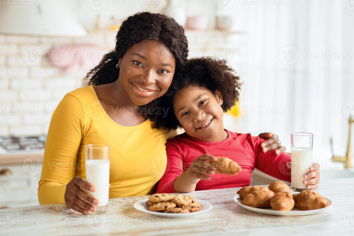 Snack Time. Happy black mother and little daughter enjoying homemade pastry and drinking milk in kitchen, having a bite with fresh baked croissants and cookies, smiling at camera, free space photo