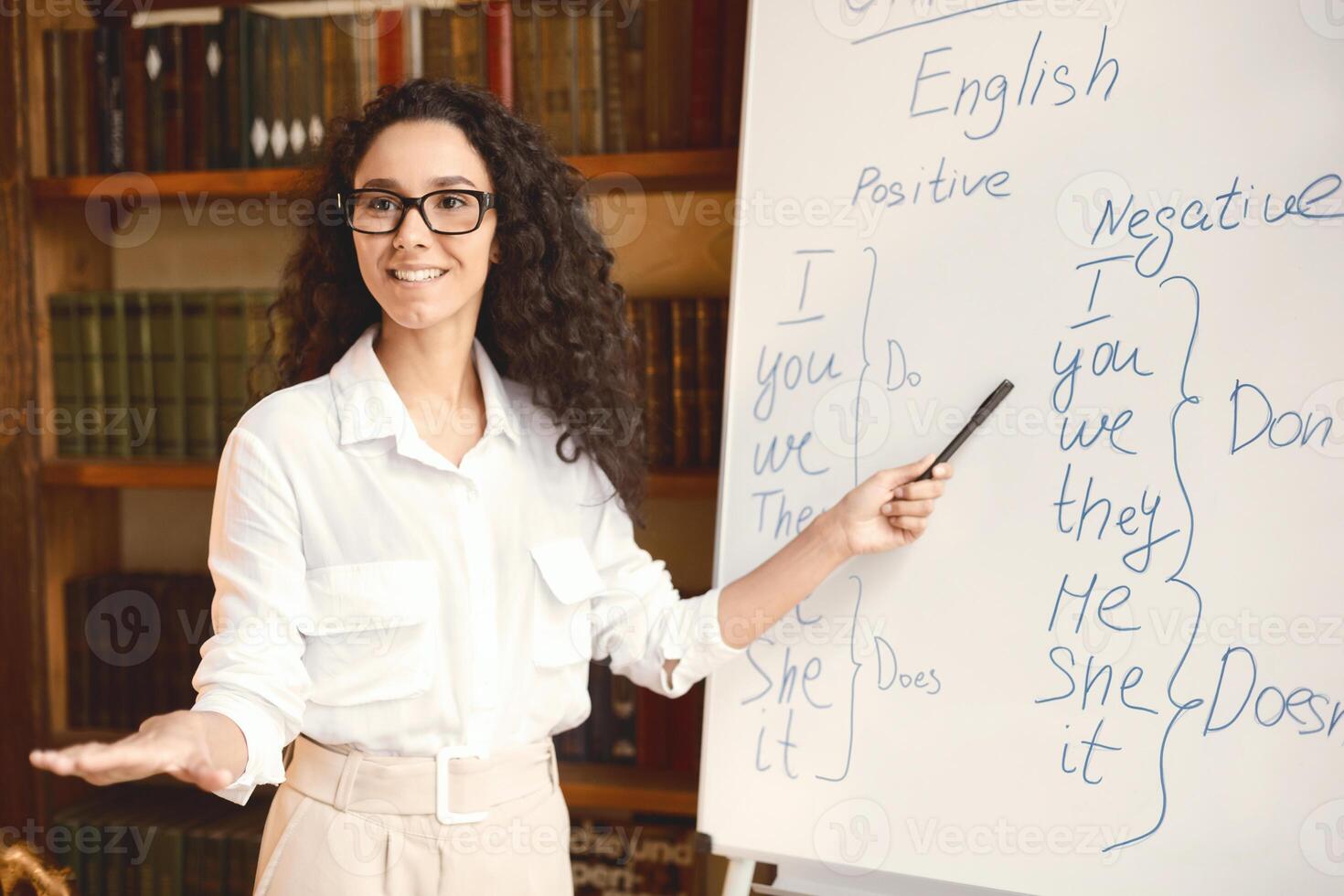 Training Concept. Portrait of cheerful positive female tutor wearing eyeglasses teaching English language, pointing at grammar rules on board with marker, looking and asking students, explaining theme photo