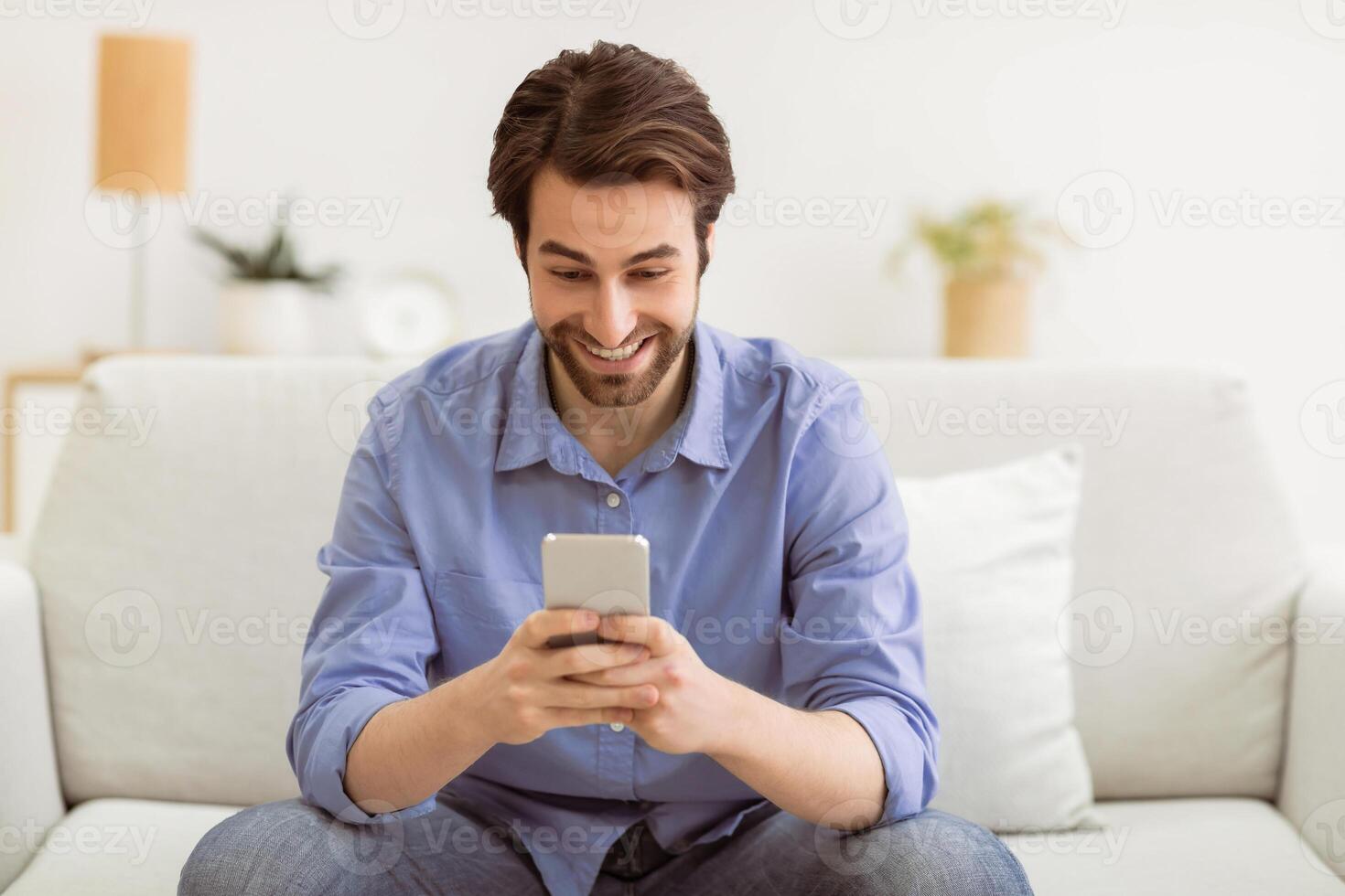 Excited Man Using Phone Networking Texting On Cellphone And Browsing Internet Sitting On Sofa At Home. New Mobile Application For Smartphone Concept. Selective Focus photo