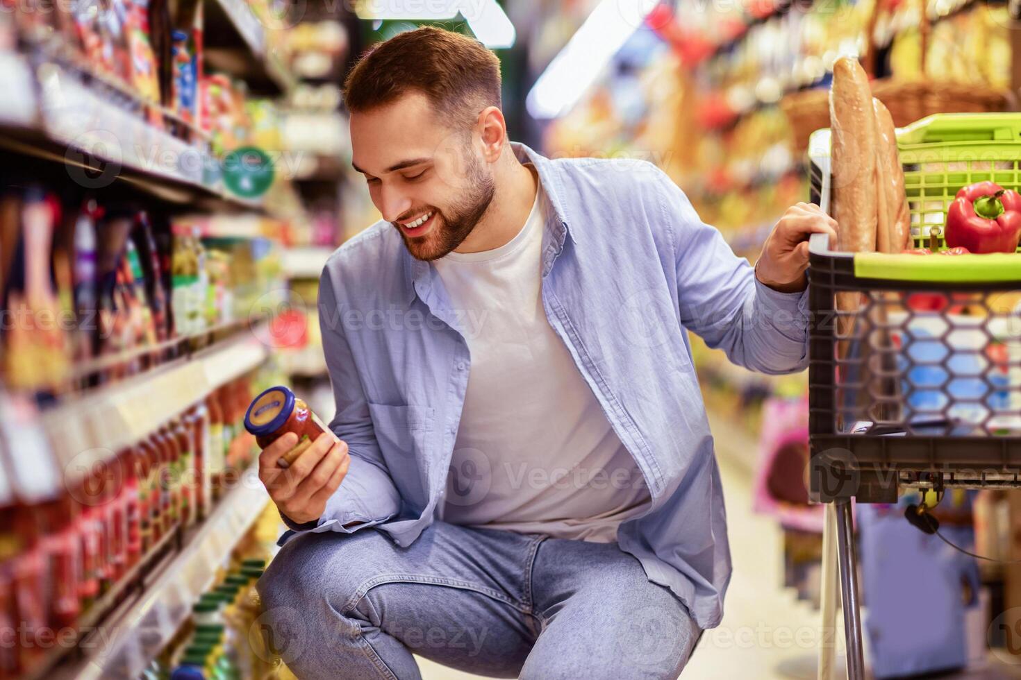 Consumption And Consumerism. Portrait Of Smiling Bearded Man With Shopping Cart In Market Buying Groceries Food Taking Products From Shelves In Store, Holding Glass Jar Of Sauce, Checking Label photo