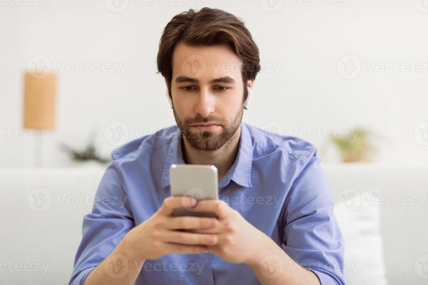 Serious Man Using Smartphone Wearing Earbuds Browsing Internet And Reading News Online Sitting On Sofa At Home. Mobile User Texting On Cellphone In Living Room. Phone Addiction Concept photo
