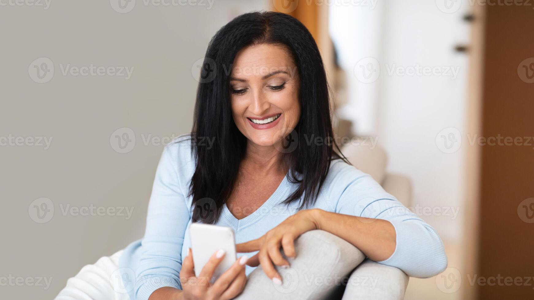 Leisure At Home. Smiling mature brunette woman using her cellphone, browsing internet, checking social media, sitting on the sofa in living room. Happy lady texting her friends, watching online photo