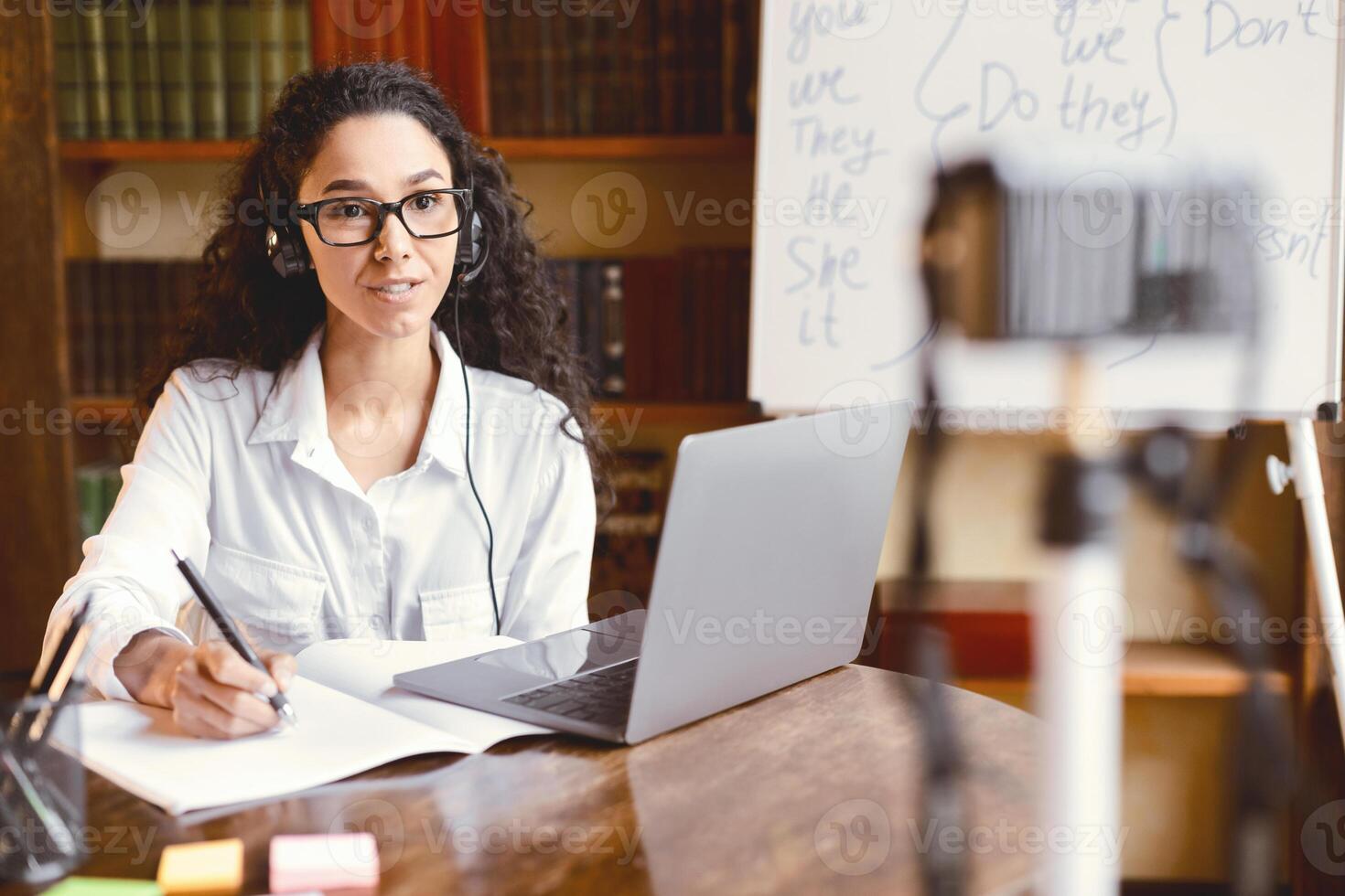 Remote Lesson. Young woman wearing eyeglasses and headset giving online class, sitting at desk, explaining new theme. Feamle teacher giving homework, filming her vlog, recording blog at camera photo