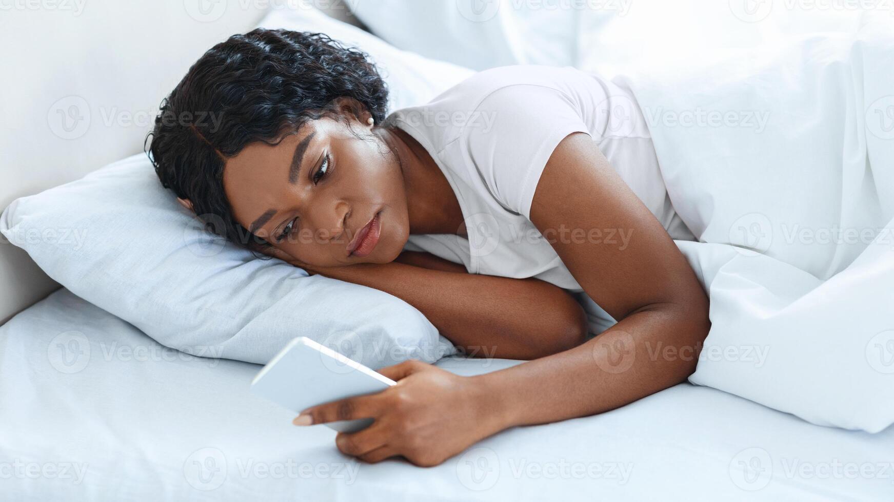 Gadget addiction, waiting for important call. Young black woman using smartphone in bed before sleeping. Frustrated african american lady checking social media while laying in bed, copy space photo