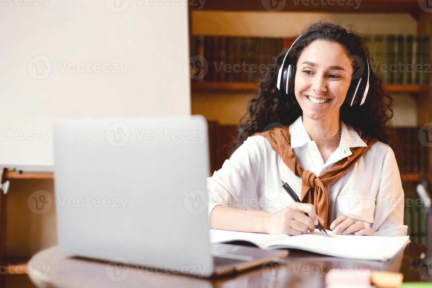 Distance Education. Smiling woman in wireless headset sitting at desk, using laptop and writing in notebook. Lady watching tutorial, lecture or webinar, studying online at home. E-learning photo