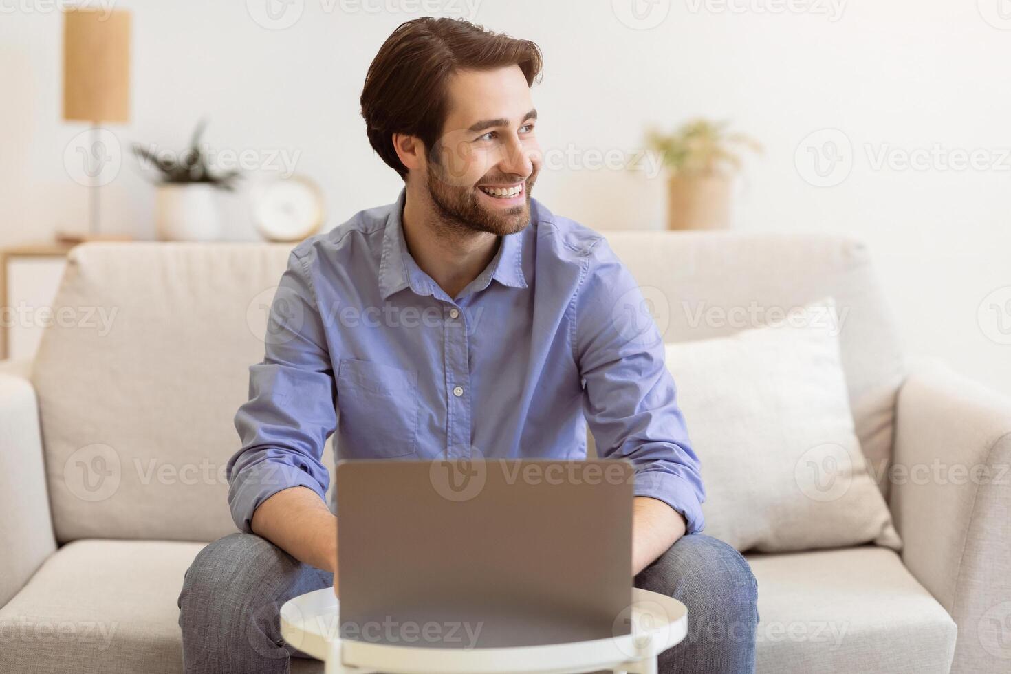 Cheerful Businessman Works On Laptop Online Smiling Looking Aside Sitting On Sofa At Home. Happy Freelancer Man Working On Computer Distantly Indoors. Freelance Job Concept photo