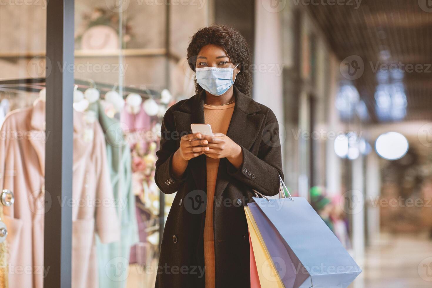 Modern gadgets for shopping and receive messages from store about sales and Black Friday. Young african american woman in protective mask holds shopping bags and uses phone to type on social media photo