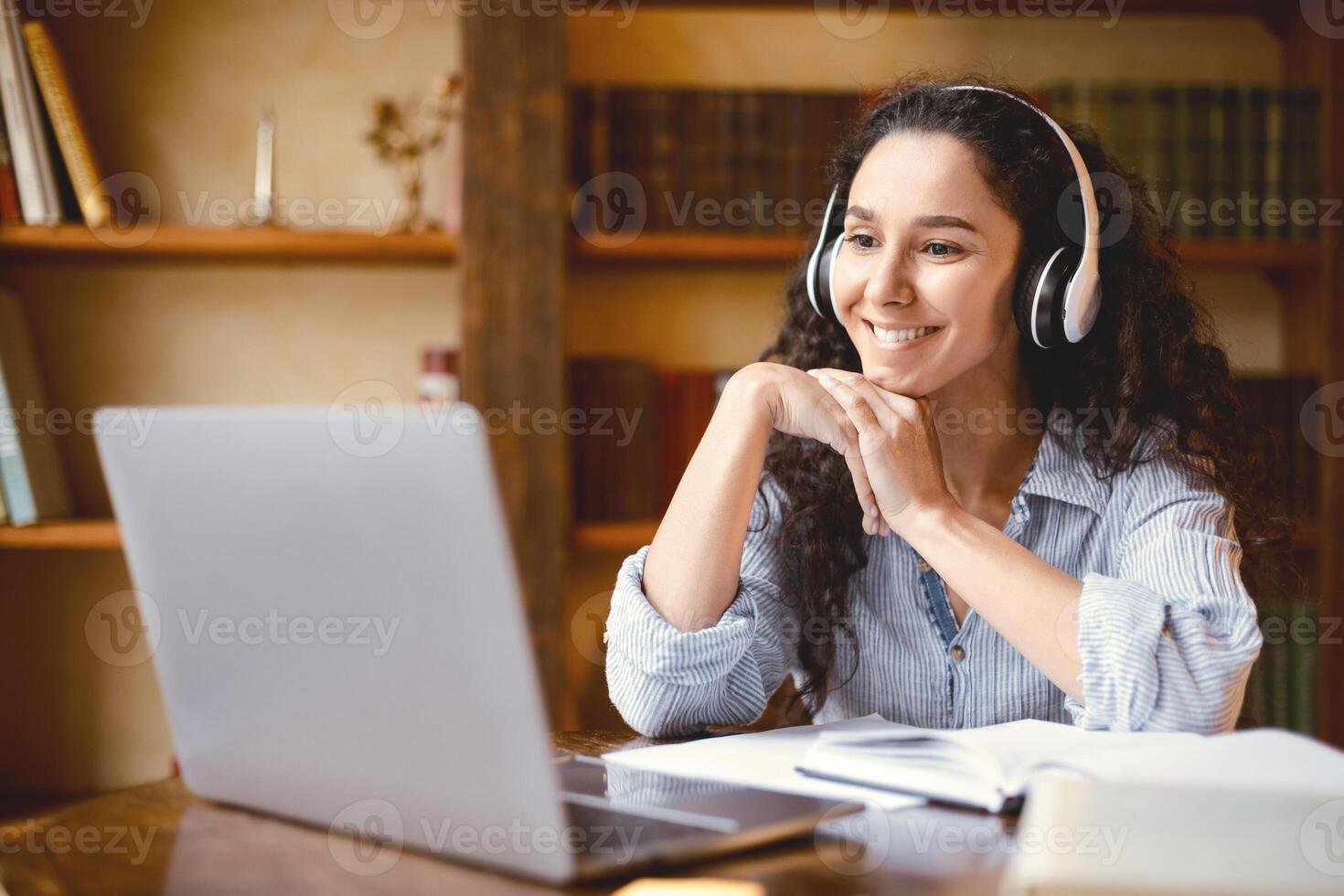 Virtual Lecture. Portrait of cheerful female student in wireless headset sitting at desk, having online class with teacher and looking at laptop computer. Smiling woman studying at home office photo